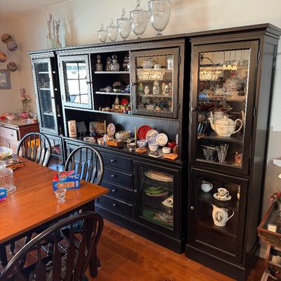 Sale Photo Thumbnail #209: Large black wooden hutch and display cabinet unit filled with various glassware, china, and decorative collectibles, shown alongside a wooden dining table and chairs.