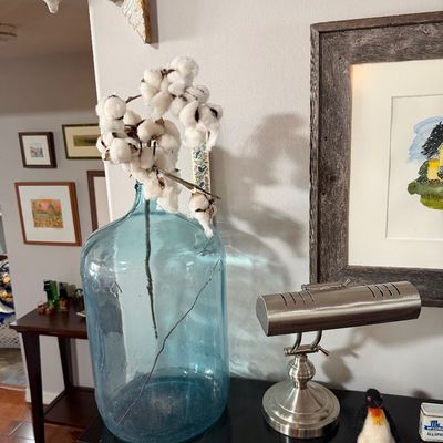 Sale Photo Thumbnail #200: Large light blue glass carboy vase holding a cotton boll branch, displayed alongside a brushed metal desk lamp and small penguin figurine.
