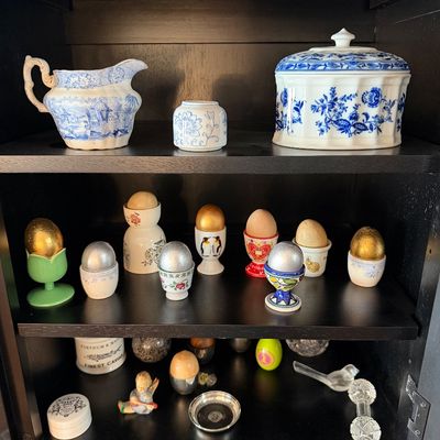 Sale Photo Thumbnail #193: Collection of vintage blue and white transferware porcelain, including a pitcher and lidded dish, displayed alongside an assortment of decorative egg cups with metallic and wooden eggs on black wooden shelves.