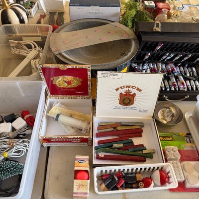 Sale Photo Thumbnail #108: Assorted garage sale items including vintage Romeo y Julieta and Punch cigar boxes filled with miscellaneous supplies, alongside a paint set, batteries, and various household goods.