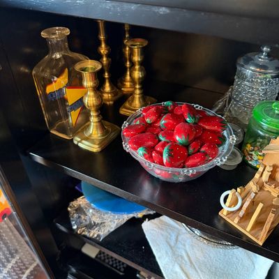 Sale Photo Thumbnail #176: Assorted decorative items on a black shelf, including brass candlesticks, a glass decanter, and a clear glass bowl filled with painted strawberry rocks.