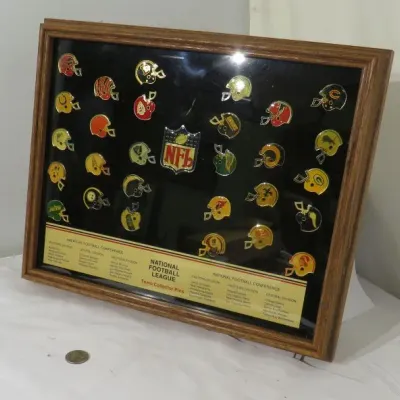 Vintage NFL team collector pins display in a wooden frame, featuring helmet-shaped pins for various franchises surrounding a central NFL...