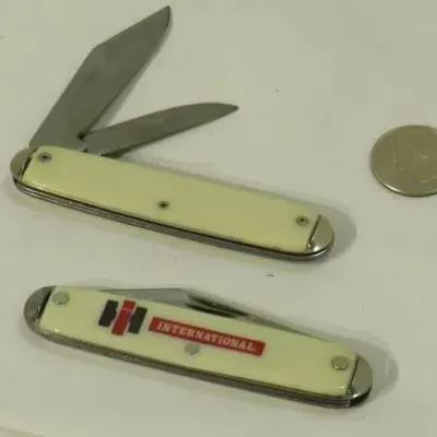 Two vintage pocket knives with cream-colored handles, including one featuring the International Harvester logo, shown next to a coin for...
