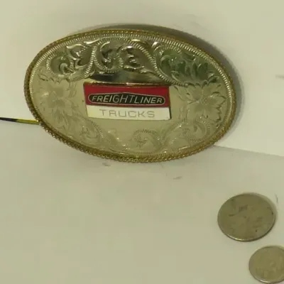 Vintage Freightliner Trucks oval belt buckle featuring a silver-tone finish with western-style floral engraving and a red and white...
