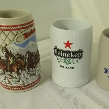 Collection of three vintage beer mugs, including a Budweiser Clydesdale holiday stein, a white ceramic Heineken mug, and a grey stoneware...