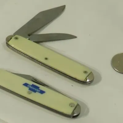 Two vintage pocket knives with cream-colored handles, one featuring a blue Chevrolet logo, shown next to a quarter for scale.