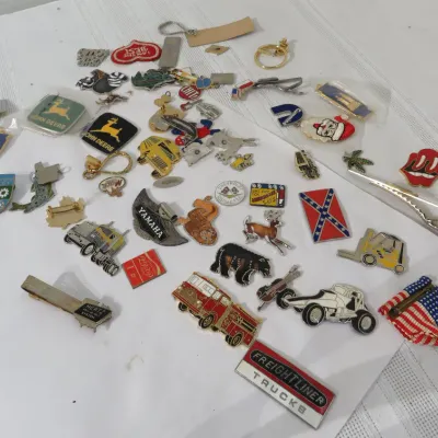 Assortment of vintage collectible pins, tie clips, and keychains, featuring brands like John Deere, CAT, Yamaha, and Freightliner.