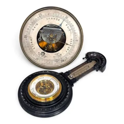 Sale Photo Thumbnail #192: Two antique aneroid barometers, including a circular brass-cased wall barometer with an open center showing the mechanism, and a German-made barometer with a thermometer mounted in a carved dark wood frame.