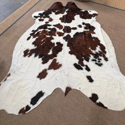 Sale Photo Thumbnail #36: Large tricolor cowhide rug featuring a natural pattern of white, brown, and black spots, displayed on a textured neutral carpet.