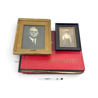 Sale Photo Thumbnail #41: Two framed antique black and white portraits, one of a man in a gold frame and one of a woman in a black frame, resting on a vintage red photograph album.