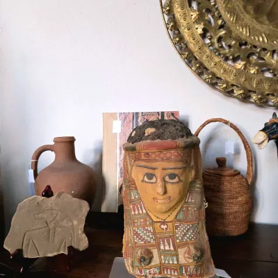 Egyptian-style painted mummy mask replica alongside terracotta pottery, carved stone relief fragment, woven basket, and brass wall plaque.
