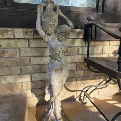 Weathered concrete garden statue depicting a woman carrying a water jug, featuring natural patina and moss growth from outdoor exposure.