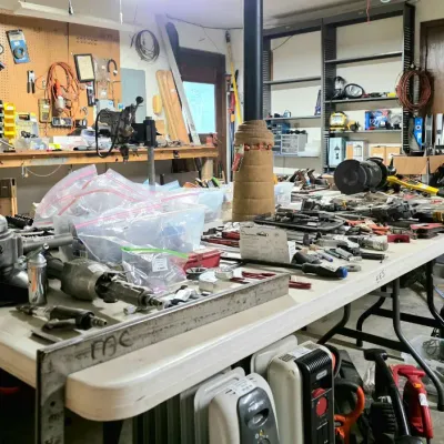 Workshop loaded with various hand tools, power tools, lawn equipment, space heaters, and hardware arranged on folding tables in a garage...