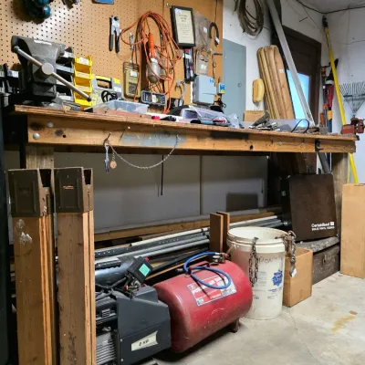 Heavy-duty wooden garage workbench equipped with a mounted bench vise, pegboard tool storage, air compressor, and portable air tank below.
