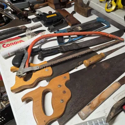 Sale Photo Thumbnail #40: Assortment of vintage hand tools, including hand saws with wooden handles, a bow saw, a machete, and various woodworking planes displayed on a table.