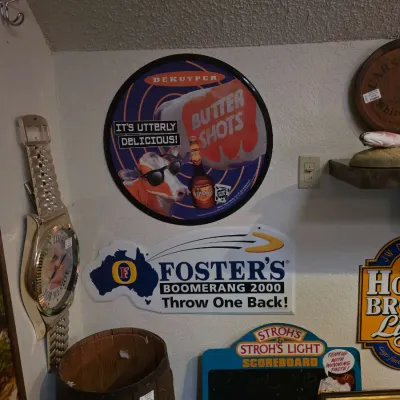 Sale Photo Thumbnail #22: Collection of vintage bar memorabilia, including a DeKuyper Buttershots round metal sign, a Foster's Boomerang 2000 tin sign, a Stroh's Light chalkboard scoreboard, and a novelty oversized wristwatch wall clock.