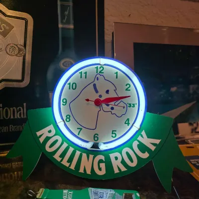 Sale Photo Thumbnail #19: Vintage Rolling Rock neon clock featuring the brand's signature horse head logo and green ribbon design, accompanied by a matching bottle opener.