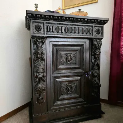Ornately carved dark wood antique cabinet featuring cherub motifs, egg-and-dart molding, and intricate paneling.