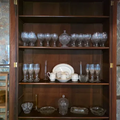 Sale Photo Thumbnail #24: Vintage wooden china cabinet filled with an assortment of cut glass stemware, serving bowls, and a small porcelain tea set.