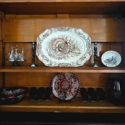 Vintage wood cabinet display featuring a large transferware turkey platter, a pair of twisted metal candlesticks, glass cruets, and...