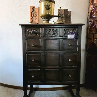 Vintage dark wood chest of drawers with carved panel detailing, topped with a brass samovar and carved wooden decorative plaques.