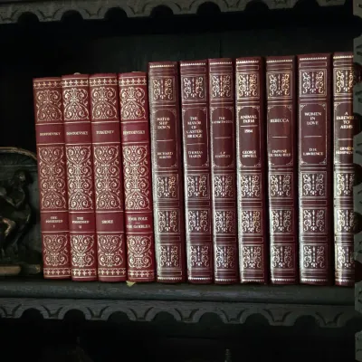 Sale Photo Thumbnail #51: Collection of classic literature books with ornate red and gold faux-leather spines, including works by Dostoevsky, George Orwell, and Daphne du Maurier, displayed on a dark wooden shelf.
