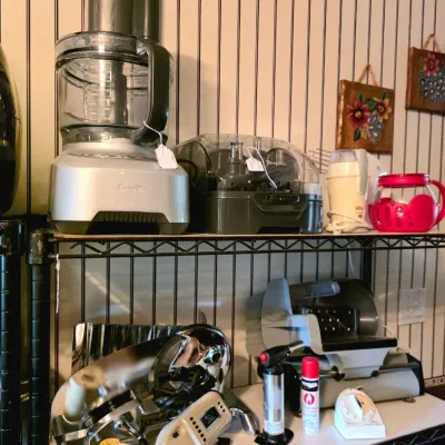 Wire shelving unit filled with countertop kitchen appliances, including a Breville food processor with accessory case, a meat slicer, and...