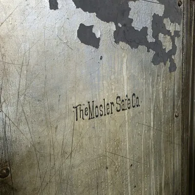 Vintage heavy-duty metal safe door featuring stenciled branding for The Mosler Safe Co., showing significant surface wear, scratches, and...