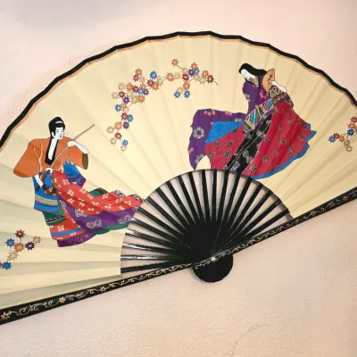 Large decorative Asian wall fan featuring painted figures in traditional dress and floral motifs on a cream-colored background with black...