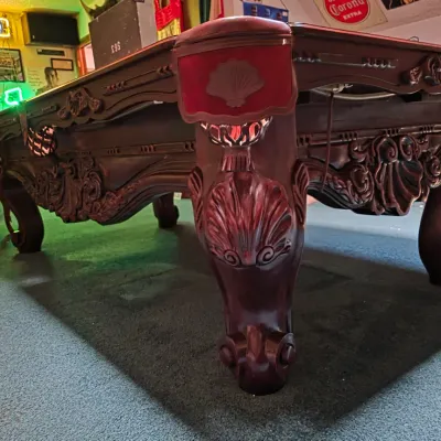 Ornate wooden pool table featuring intricate shell and scroll carvings on the legs and apron, finished in a rich dark cherry stain.