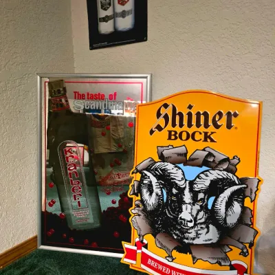 Collection of bar memorabilia, featuring a yellow Shiner Bock metal sign with a ram graphic and a framed Kränberi Vodka mirror sign.