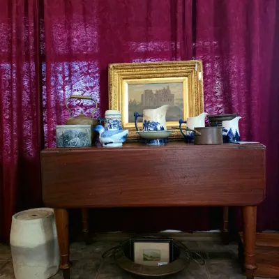 Antique drop-leaf wooden table displayed with blue and white porcelain pitchers, a framed landscape oil painting, a copper kettle, and...