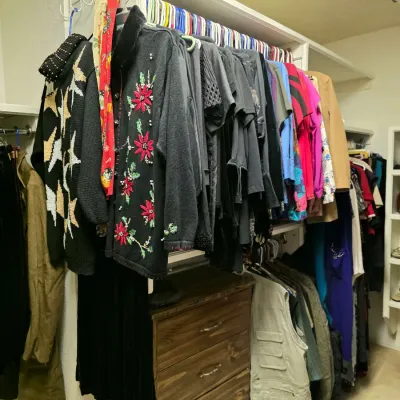 Walk-in closet filled with vintage women's clothing, including embellished cardigans, vests, and colorful tops, alongside a wooden...