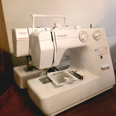Pair of white Singer portable electric sewing machines, including a modern free-arm model with stitch dials.