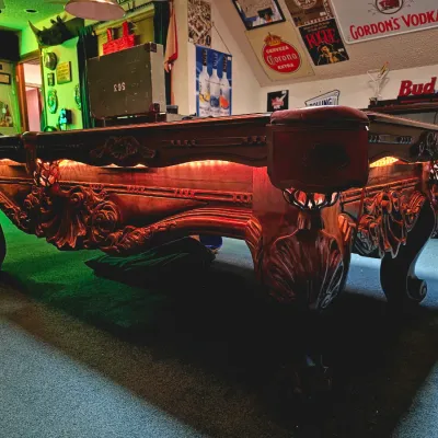 Ornate carved wood pool table with ball-and-claw feet, leather drop pockets, and custom under-rail lighting.