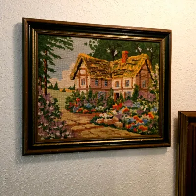 Vintage framed needlepoint artwork depicting a cottage with a thatched roof and a colorful flower garden, housed in a dark wood frame.