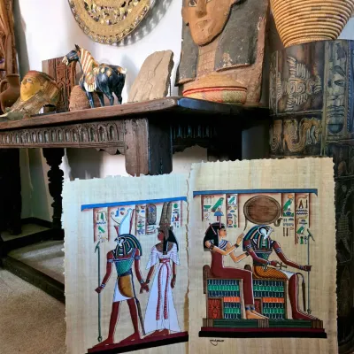 Sale Photo Thumbnail #48: Two Egyptian papyrus paintings depicting deities, displayed alongside an assortment of Egyptian-revival and global decor, including a carved wooden console table, a large wall mask, and a brass charger.