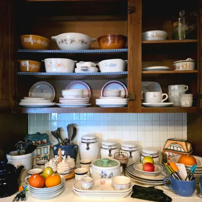A collection of vintage kitchenware and tableware displayed in and under wooden cabinets, including Pyrex casserole dishes, CorningWare,...
