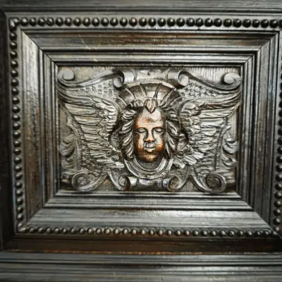 Intricately carved dark wood furniture panel featuring a winged cherub motif surrounded by beaded trim and molded framing.