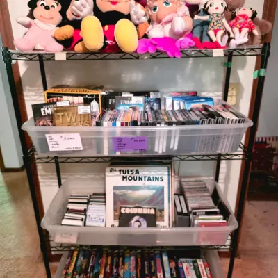 Wire shelving unit filled with plush character toys, clear plastic bins of DVDs, CDs, audio cassette tapes, and vinyl record albums.