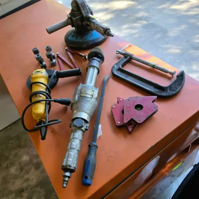 Sale Photo Thumbnail #34: Assorted power and hand tools, including a yellow angle grinder, a pneumatic die grinder, a C-clamp, magnetic welding holders, and a pry bar, displayed on an orange workbench.