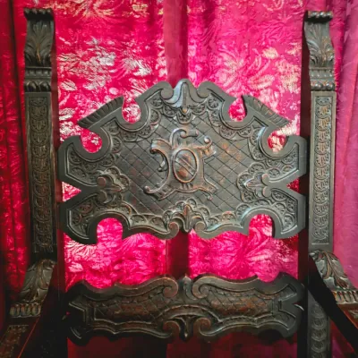 Sale Photo Thumbnail #16: Ornate Gothic Revival style carved wooden armchair featuring a high back with a central crest and intricate relief details, set against a red damask background.