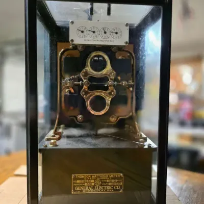 Vintage General Electric Thomson Watthour Meter enclosed in a glass and metal display case, featuring intricate brass internal components...
