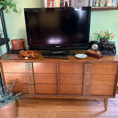 Mid-century modern six-drawer dresser with carved wooden pulls and tapered legs, supporting a Samsung flat-screen television and various...