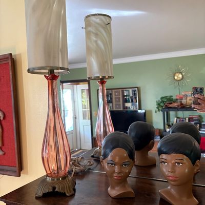 Mid-century modern pink glass table lamp with a tall cylindrical shade, displayed alongside a vintage ceramic female bust on a wooden...