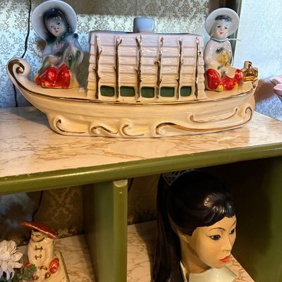 Vintage mid-century ceramic TV lamp shaped like a house boat with Asian figures, shown alongside a ceramic lady head vase on a green shelf.
