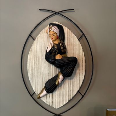 Sale Photo Thumbnail #174: Mid-century modern wall plaque featuring a chalkware dancer in a black and gold outfit, mounted on a textured oval backing with a black wire frame.