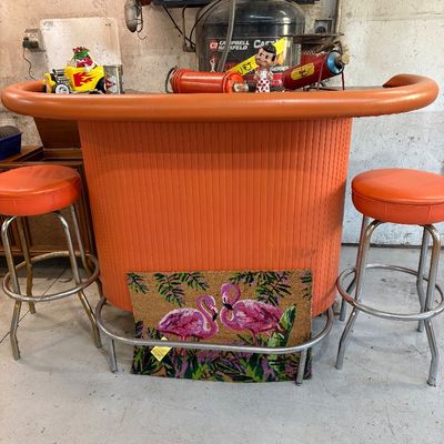 Mid-century modern home bar with ribbed orange paneling and a padded orange armrest, accompanied by two matching orange vinyl barstools...