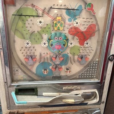 Sale Photo Thumbnail #274: Vintage Nishijin Deluxe Super pachinko machine featuring a colorful playfield with a central mask motif, showing some wear and dust from age.