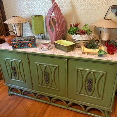 Mid-century olive green painted credenza with decorative door panels and arched base, styled with vintage table lamps, a pink ceramic...